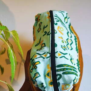 May include: A small, rectangular zippered pouch with a blue and white fabric exterior featuring a geometric pattern in yellow and green. The pouch is lined with a brown fabric.