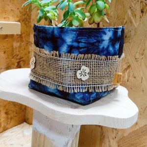 Puede incluir: Una maceta de tela azul y blanca con estampado tie-dye, con un borde de arpillera y detalles de flores de madera. La maceta está llena de una planta suculenta y se encuentra sobre un soporte de madera blanco.