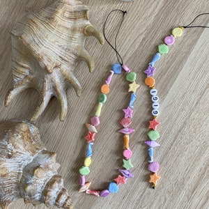 May include: Two colorful beaded necklaces with a seashell theme. One necklace has the name "GIGI" spelled out in white beads. The necklaces are on a wooden surface with two seashells.
