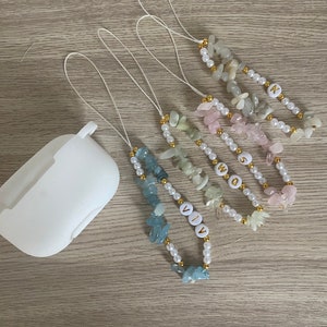 Puede incluir: Cinco charms diferentes para la correa de Airpod con cuentas de perlas blancas y piedras de varios colores. Cada charm tiene un nombre escrito con letras doradas: VIV, SWQ, AIVIA, N.