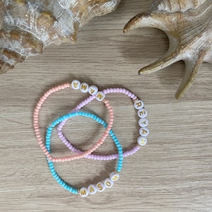 May include: Three beaded bracelets with the names "MASON", "REIAH", and "MANA" spelled out in gold letters on white beads. The bracelets are layered on a light brown wooden surface.