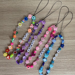 Può includere: Una collezione di ciondoli per telefono con perline di vari colori. Ogni ciondolo presenta un cordino nero e un design unico con perline di lettere che compongono nomi. I ciondoli includono frutta, fiori e altre perline decorative.