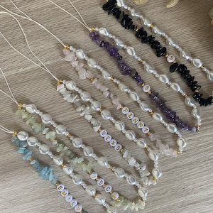 Puede incluir: Una colección de collares con cuentas de perlas blancas y diferentes chips de piedras preciosas de colores. Cada collar presenta un dije de nombre personalizado, incluyendo "Emily", "Jess", "Bexs", "Penny" y "S".