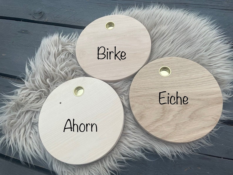 May include: Three round wooden boards with brass eyelets. The boards are labeled "Birke", "Eiche", and "Ahorn" in black script. The boards are arranged on a fluffy, light-colored surface.