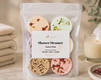 May include: A clear plastic pouch containing a variety pack of round shower steamers in shades of peach, light green, purple, tan, and pink, each topped with dried botanicals. The label reads Shower Steamer Variety Pack, Net wt 1.5 oz | 42 g each, Breathe, Relax, Unwind.