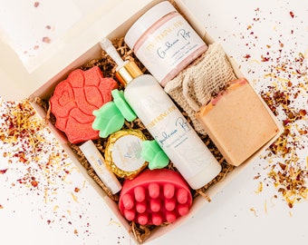 May include: A gift box filled with bath and body products, including a rose-shaped bath bomb, a green leaf-shaped soap, a pink bath scrub, a white lotion bottle, a pink sugar scrub jar, a bar of soap, and a lip balm. The box is lined with brown paper and the products are arranged in a visually appealing way.