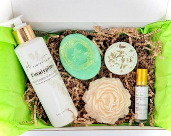 May include: A gift box filled with eucalyptus-themed bath and body products. The box contains a green bath bomb, a green soap bar, a white bottle of eucalyptus body lotion, a white bottle of eucalyptus aromatherapy oil, and a beige soap bar shaped like a flower.