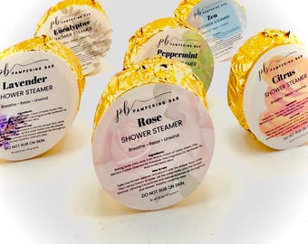 May include: Six round shower steamers wrapped in gold foil. Each steamer has a different scent: Lavender, Eucalyptus, Peppermint, Zen, Rose, and Citrus. The steamers are labeled with the scent and the words "Shower Steamer" and "Breathe Relax Unwind".