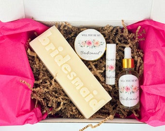 May include: A gift box filled with items for a bridesmaid proposal. The box contains a bar of soap with the word "bridesmaid" embossed on it, a small round container with a floral design and the text "Will you be my bridesmaid?", a lip balm with a floral design and the text "Will you be my bridesmaid", and a small bottle of oil with a floral design and the text "Will you be my bridesmaid?"