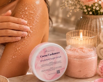 May include: A round container of Cocoa Cashmere emulsified sugar scrub with a pink label, next to an open jar of pink exfoliating scrub. The label text reads Cocoa Cashmere Emulsified Sugar Scrub, Net wt 5 oz / 142 g, with ingredients and instructions.