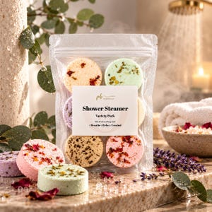 May include: A clear plastic pouch containing a variety pack of round shower steamers. The label reads Shower Steamer, Variety Pack, Net wt 1.5 oz | 42 g each, Breathe - Relax - Unwind. The steamers feature dried flower petals and herbs.