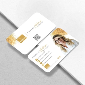 May include: Two white business cards with rounded corners. One card features the name "TIFFANY Anderson REALTOR" in gold script, a QR code, and the "RealLiving" logo. The other card has a photo of a woman and contact information.