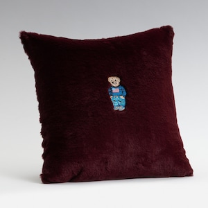May include: A burgundy faux fur cushion with an embroidered image of a brown bear wearing a blue shirt with a red, white and blue flag on the front.