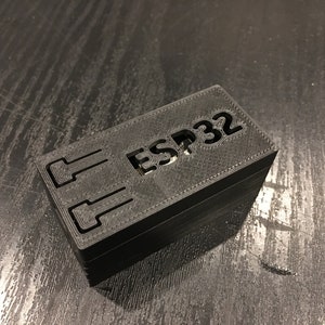 May include: A black 3D printed case with the text "ESP32" printed on the top. The case has two cutouts on the side.
