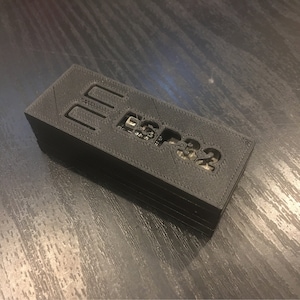 May include: A black 3D printed case with the text "ESP32" printed on the top. The case has three rectangular openings on the top.