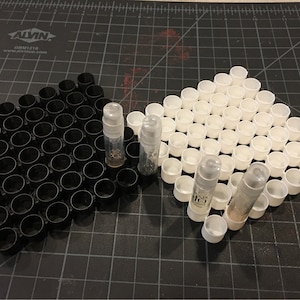 May include: A black and white grid of small plastic containers with three clear glass bottles filled with a white substance. The bottles are standing upright in the containers.
