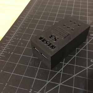 May include: A black plastic case with the text "CS Dustin" printed on the front. The case has several small holes and a rectangular slot on the top.