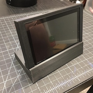 May include: A black 3D printed stand for a tablet or phone. The stand has a raised back and a flat base. The stand is on a gray grid surface.