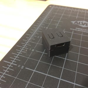 May include: A black plastic box with three rectangular openings on the top and a micro USB port on the side. The box is sitting on a black cutting mat.