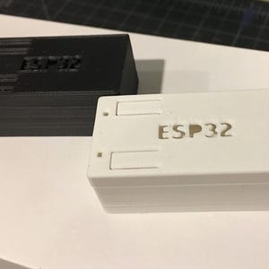 May include: Two rectangular ESP32 electronic component enclosures. One is black, the other is white. Both have the text "ESP32" on the top. The black enclosure is stacked, and the white enclosure has two rectangular cutouts.