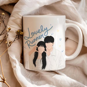 May include: A white ceramic mug featuring a blue script text that reads Lovely Runner above a stylized illustration of two people making a heart shape with their hands. The design includes small yellow and blue heart and star accents.