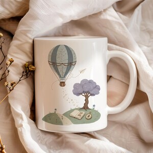 Könnte beinhalten: Weiße Keramik-Tasse mit einer skurrilen Illustration eines Heißluftballons, eines Baumes und eines offenen Buches. Der Ballon ist blau und grau, mit einem braunen Korb. Der Baum hat ein lilafarbenes Blätterdach. Ein Papierflieger und Sterne sind ebenfalls abgebildet.