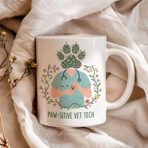 Könnte beinhalten: Weiße Keramiktasse mit einem skurrilen Design. Die Tasse zeigt einen Tierarzt in OP-Kleidung, der einen Pfotenabdruck hält, mit einem Pfotenabdruckkopf und floralen Akzenten. Der Text "PAW-SITIVE VET TECH" ist auf der Vorderseite aufgedruckt.
