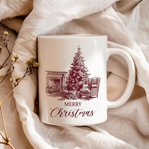 Op de afbeelding: Witte keramische mok met een illustratie van een kerstboom, open haard, fauteuil en cadeaus. De woorden "Merry Christmas" staan eronder. De mok heeft een groot handvat.