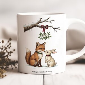 Könnte beinhalten: Weiße Keramik-Tasse mit einer Aquarell-Illustration eines Fuchses und eines Kaninchens unter einem Mistelzweig. Der Fuchs ist braun und das Kaninchen ist weiß. Die Tasse trägt unten den Text "Midnight Mistletoe Meeting".