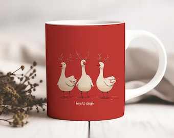 Op de afbeelding: Een witte keramische mok met een rode achtergrond. De mok toont drie cartoon ganzen met geweien, met de tekst "here to sleigh". De mok heeft een wit handvat en staat tegen een wazige achtergrond.