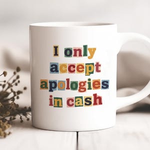 May include: White ceramic mug with the text "I only accept apologies in cash" in colourful block letters. The mug has a curved handle. The background is blurred, with a sprig of dried flowers.