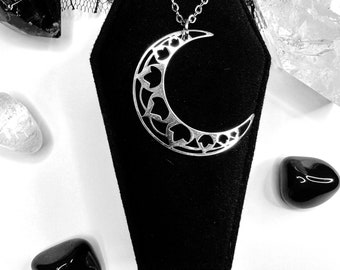 May include: A silver crescent moon necklace with a delicate heart design. The necklace is displayed on a black velvet coffin-shaped box.