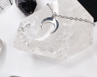 May include: A silver crescent moon pendant necklace on a silver chain, resting on a clear quartz crystal. The necklace is surrounded by other crystals, including a black crystal and a clear quartz crystal.