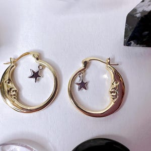 May include: A pair of gold-coloured hoop earrings. Each earring features a crescent moon design with a face and a dangling star charm. The earrings are set against a white background, with crystals in the background.