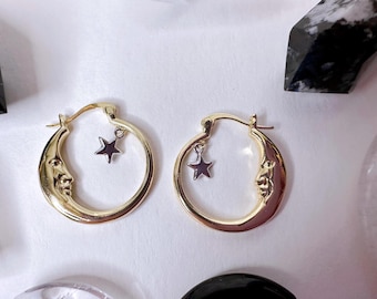 Könnte beinhalten: Ein Paar goldfarbene Creolen. Jeder Ohrring hat ein Mond-Design mit Gesicht und einen baumelnden Stern-Anhänger. Die Ohrringe sind vor einem weißen Hintergrund mit Kristallen im Hintergrund platziert.