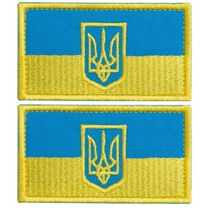 Op de afbeelding: Twee geborduurde patches met het ontwerp van de Oekraïense vlag. Elke patch heeft een blauw bovenste gedeelte en een geel onderste gedeelte, met een geel geborduurd drietandembleem in het midden. De patches zijn rechthoekig met gele randen.