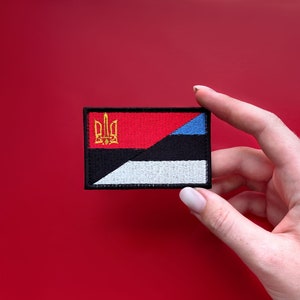 Könnte beinhalten: Gestickter Aufnäher mit Flaggenmotiv. Er zeigt rote, schwarze, weiße und blaue Abschnitte. Ein goldenes Emblem befindet sich auf rotem Hintergrund in der oberen linken Ecke. Der Aufnäher ist rechteckig mit schwarzem Rand.