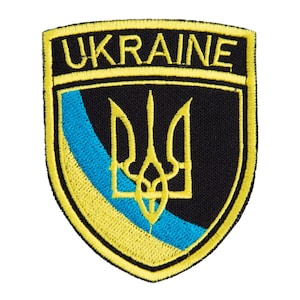 Puede incluir: Parche bordado con la palabra "UKRAINE" en dorado sobre un escudo negro. El escudo muestra un emblema de tridente dorado y una franja diagonal azul y amarilla, los colores de la bandera ucraniana.