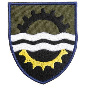 Op de afbeelding: Geborduurde patch in schildvorm. Het ontwerp bevat een zwart tandwiel, witte golvende lijnen en een geel tandwiel op een olijfgroene achtergrond. De patch heeft een donkerblauwe rand.