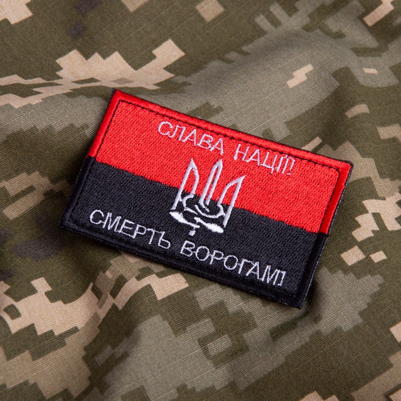 May include: A rectangular patch with a red and black design, featuring white text and a trident symbol. The patch is set against a camouflage background.