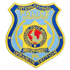 May include: Embroidered patch featuring the International Police Association logo. The shield-shaped patch has a light blue background with a gold star and a globe in the center. The text "INTERNATIONAL POLICE ASSOCIATION" and "UKRAINIAN SECTION IPA" are visible.