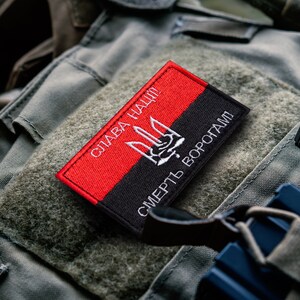 May include: A rectangular patch with a red and black design. The red section features white text that reads "Слава Нації!" and a white trident symbol. The black section has white text that reads "Смерть Ворогам!"