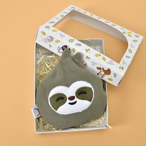 May include: A green sloth-shaped plush toy with a smiling face, presented in a white gift box with a woodland animal design. The box is open, revealing the toy nestled on straw. A small rainbow tag is attached.
