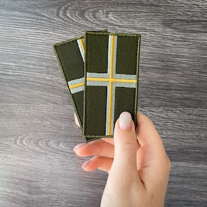 May include: Two olive green rectangular patches with a cross design. The cross is made of a light gray and yellow stripe. The patches are embroidered with a dark green border.