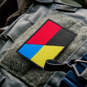 May include: A rectangular patch with a color block design. The patch is divided into four sections: red, black, blue, and yellow. The patch is on a green fabric background.