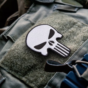 May include: White and black skull patch with a stitched design. The patch features a skull with black eye sockets and a vertical line design. The patch is placed on a green fabric background.