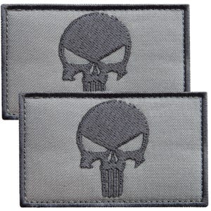 May include: Two rectangular patches featuring a dark gray embroidered skull design on a light gray fabric background. Each patch has a dark gray stitched border and a stylized skull graphic with hollow eyes and a vertical-lined nose area.