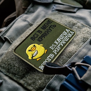 May include: An olive green embroidered patch featuring a smiling yellow chick with the text "ВСІ В МЕНЕ КРЯКНУТЬ" and "PS.КАЧЕЧКА ВОРОГІВ РОЗ'ЯРУЧЕЧКА" in white. The patch is rectangular and sits on a textured, olive green surface.