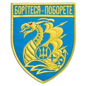 Puede incluir: Parche bordado con fondo azul y diseño de dragón amarillo. El parche presenta la frase ucraniana "БОРІТЕСЯ-ПОБОРЕТЕ" en letras amarillas. El dragón sostiene una lanza y un tridente.
