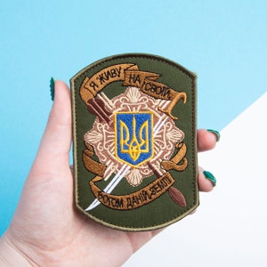 May include: An olive green embroidered patch featuring a gold emblem with a blue and yellow crest. The patch has text in Ukrainian. The patch is rectangular with rounded corners.